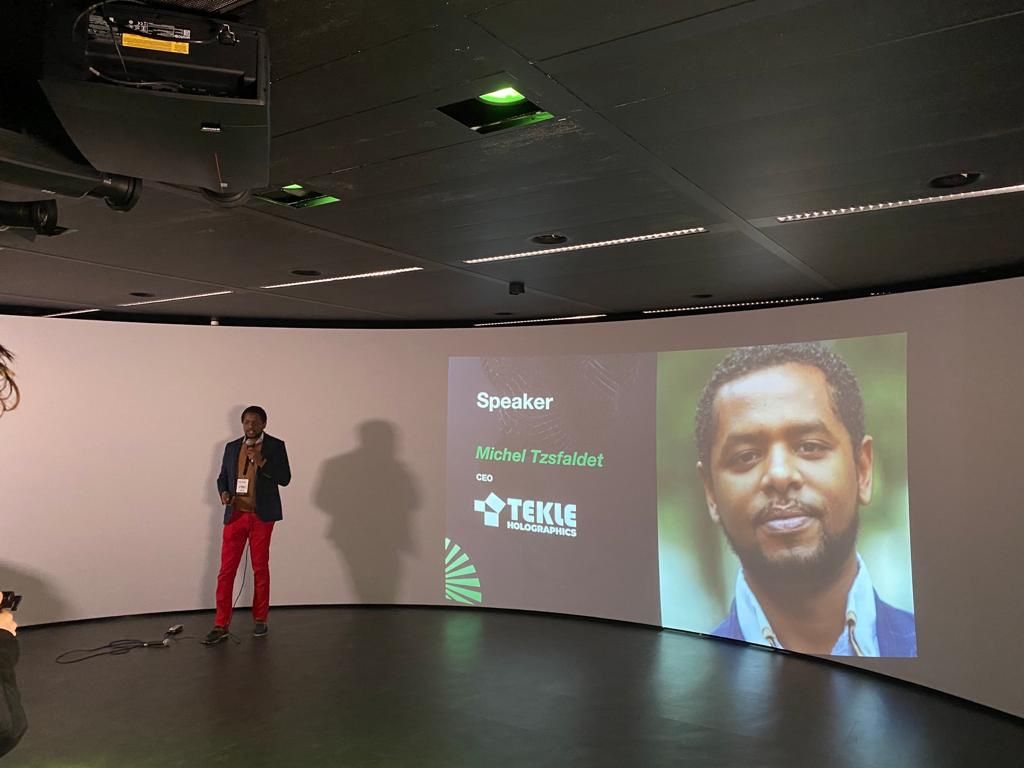 Michel Tzsfaldet op het podium tijdens een keynote in Amsterdam, met zijn naam en bedrijf op het scherm achter hem.