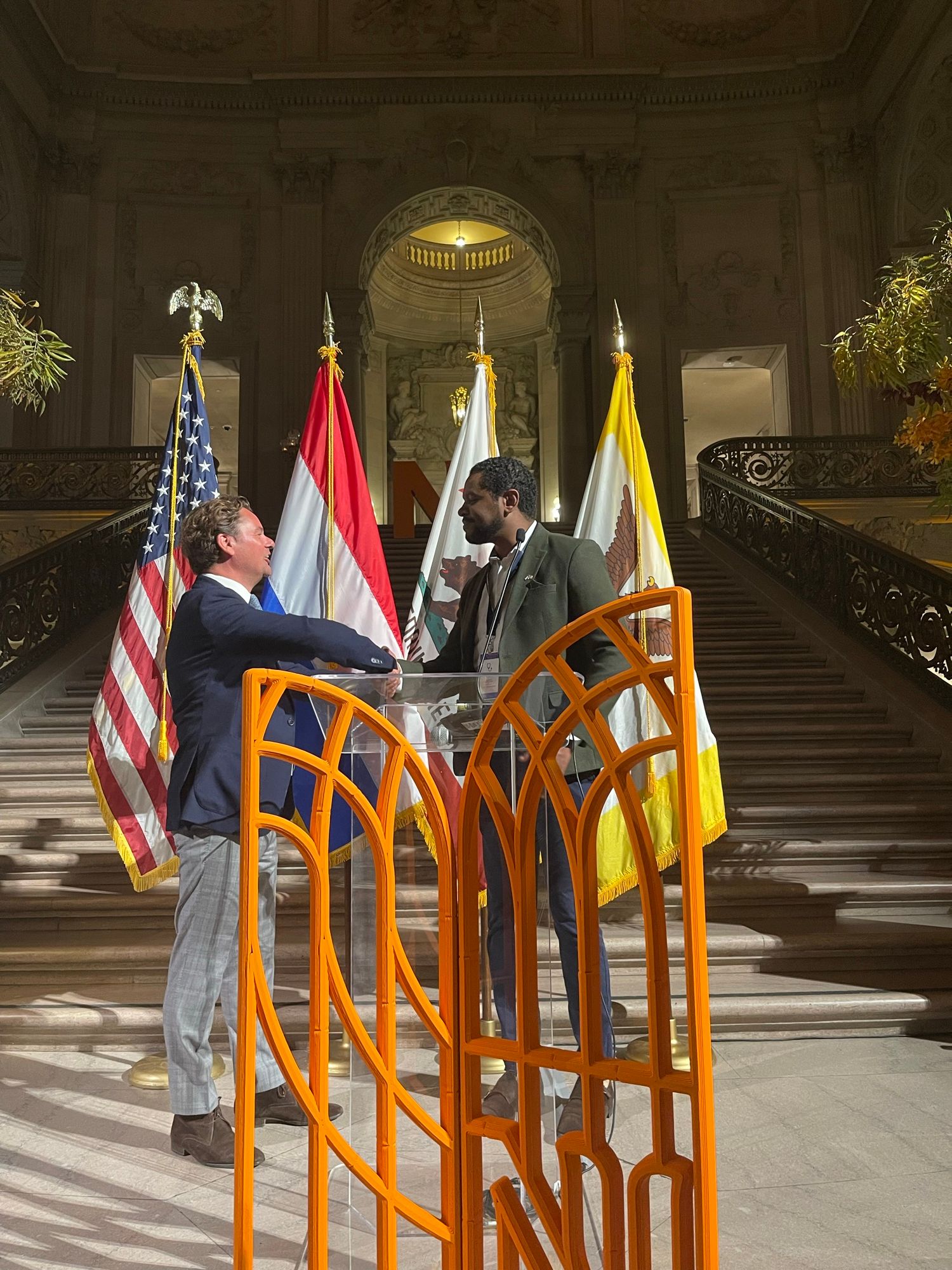 Michel Tzsfaldet beim Händeschütteln während der niederländischen Wirtschaftsmission nach Kalifornien, in der San Francisco City Hall neben US-amerikanischer und niederländischer Flagge.